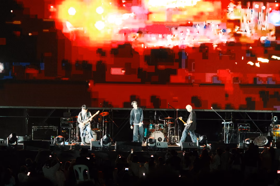 Band on stage under a screen of red and white pixelated blocks, three musicians lit in white light.