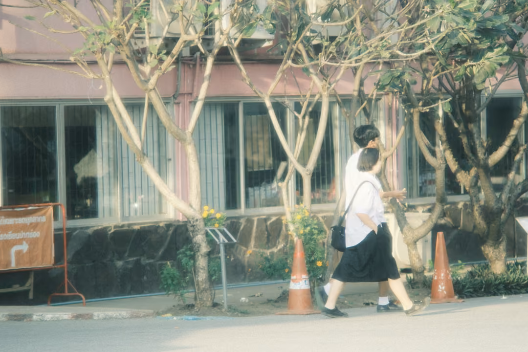 Two students in white shirts and dark skirts walk past frangipani trees and orange traffic cones beside a building.