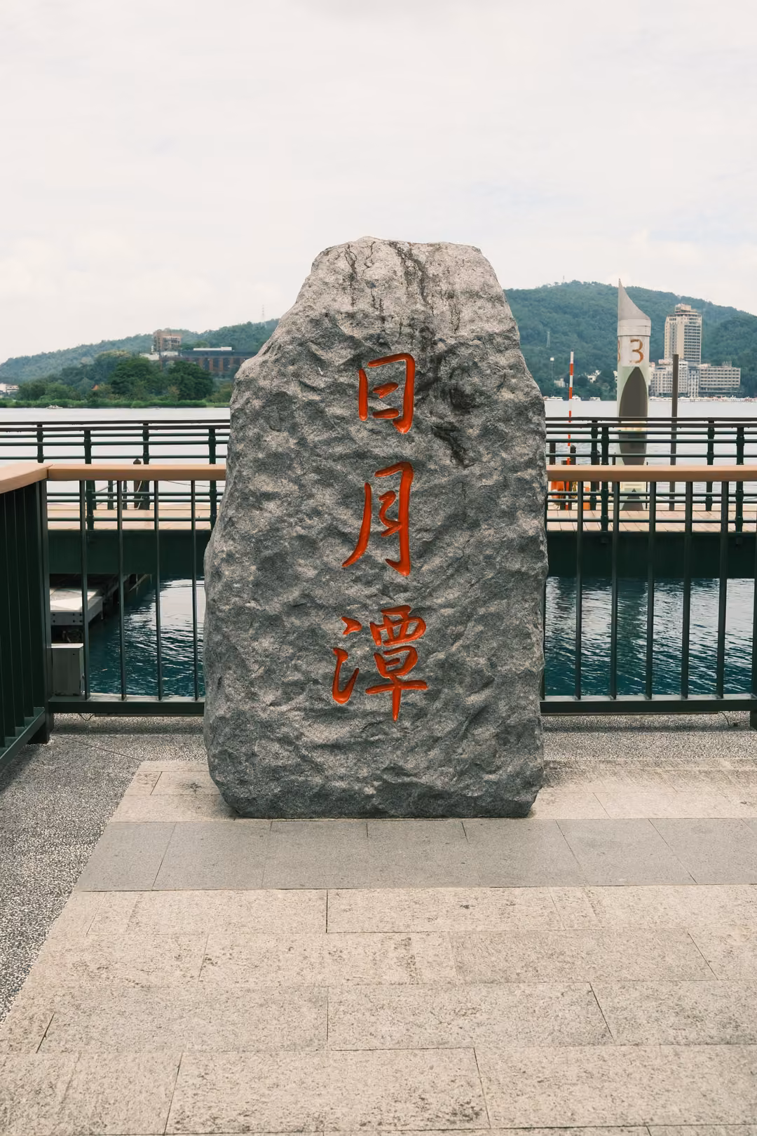 Tall grey stone carved with red characters standing alone on a lakeside terrace with a green railing