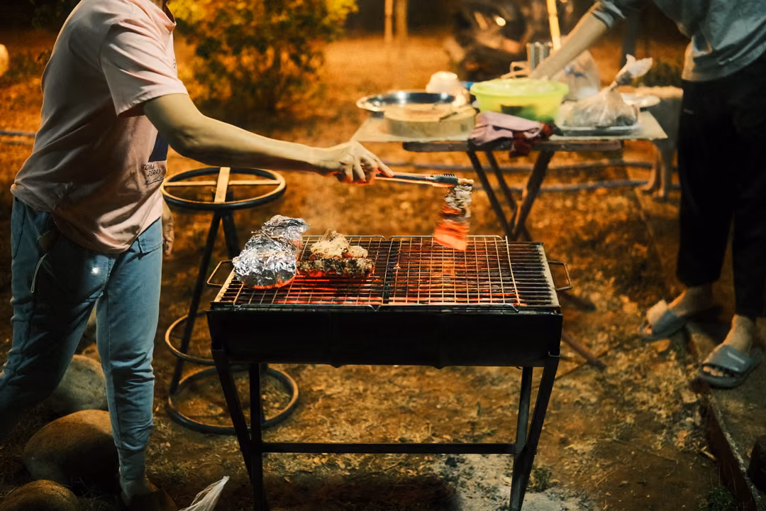 Person in jeans turning meat with tongs on a metal grill, foil packets on the grate, table of food behind