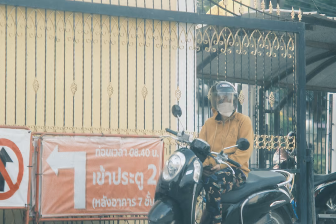 A helmeted rider in a mask and orange shirt on a scooter by an ornate gate, beside a red banner with a white arrow.