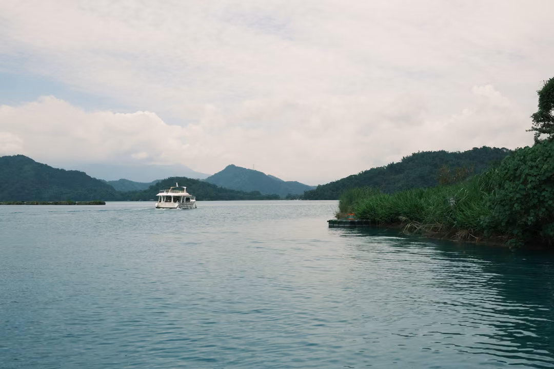 Channel of calm water between green shores with a white boat approaching and blue mountains beyond