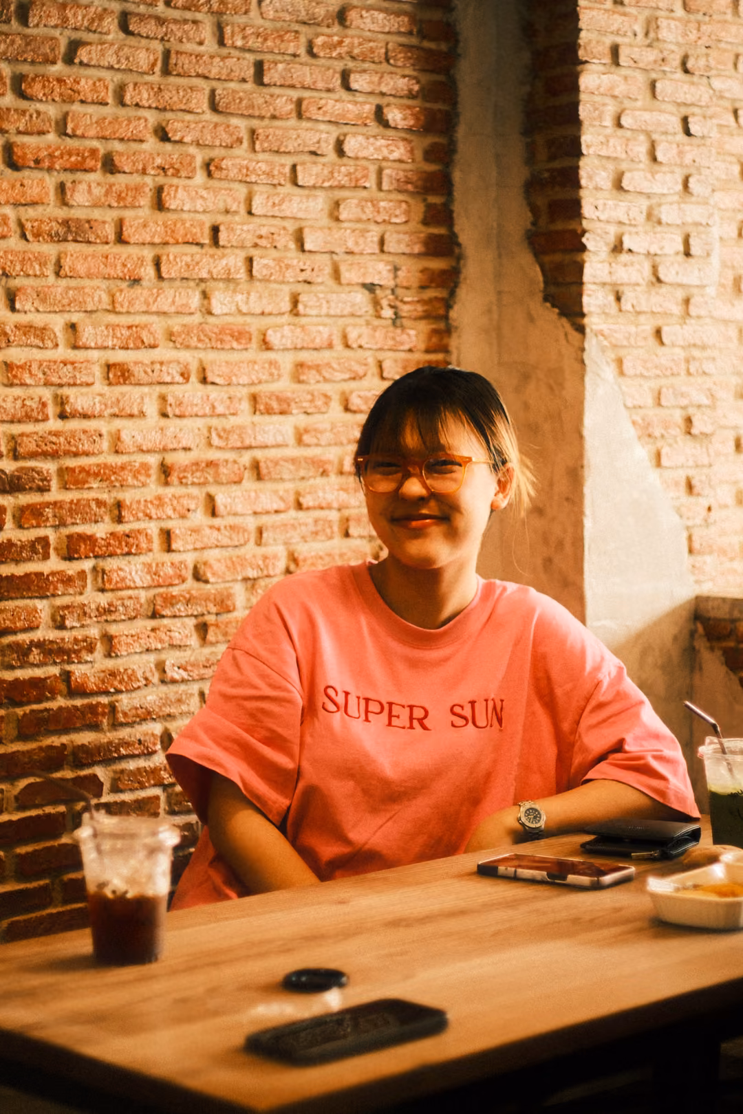 A woman in glasses and a pink SUPER SUN T-shirt smiles at a wooden table with an iced drink, brick wall behind
