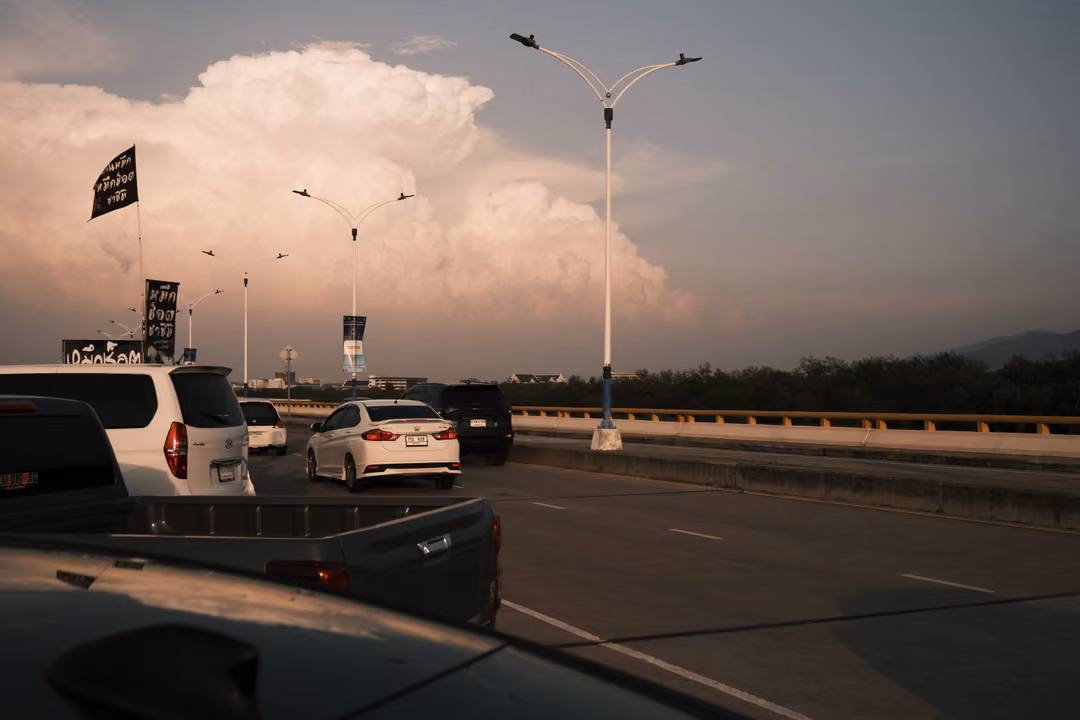 Parked cars on a causeway under tall street lamps and black flags, a towering cloud lit pink