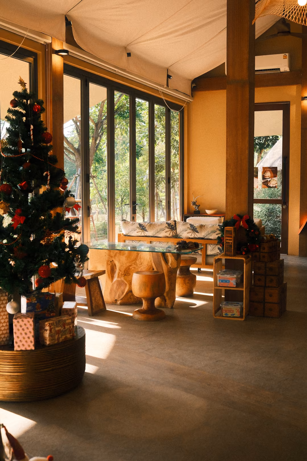 Bright interior with a decorated Christmas tree, wooden pillars, glass walls and sunlit floor under a canvas ceiling