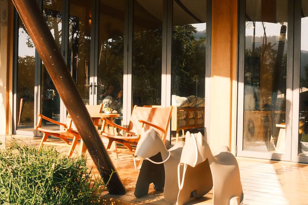 Sunlit deck with white horse-shaped seats and wooden chairs beside tall glass doors, a dark pole slanting across