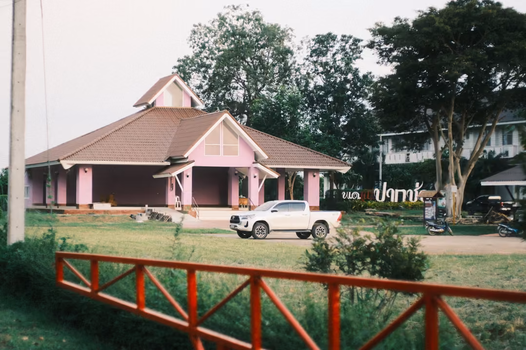 Pink-roofed building with a white pickup in front, red railing and trees, pale sky
