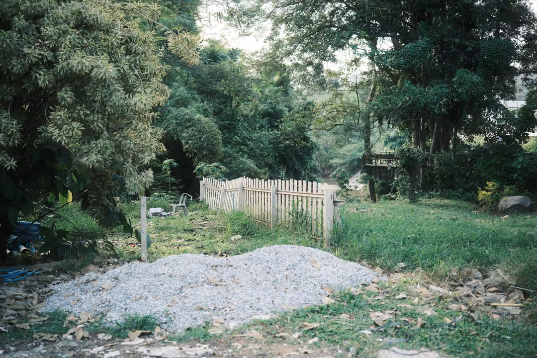 Pile of grey gravel beside a white picket fence and overgrown green vegetation