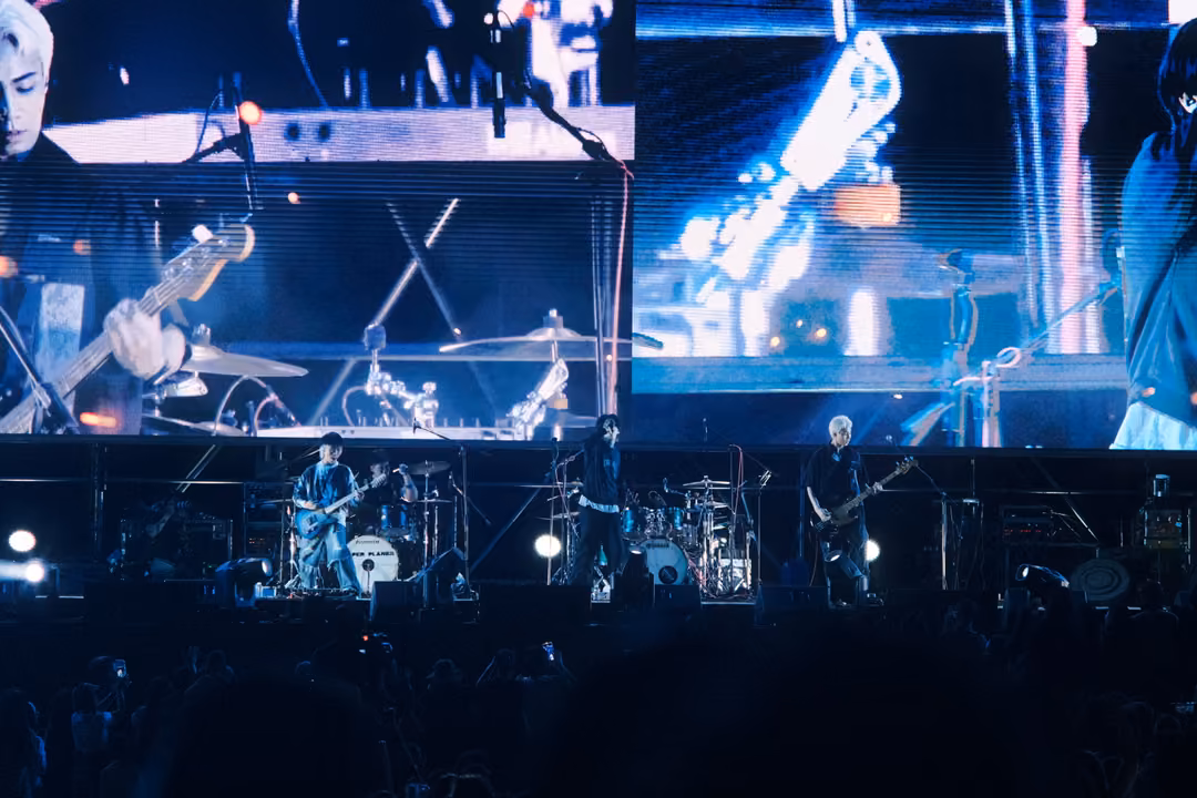 Screen split between a guitarist's hands on the strings and a blurred guitar headstock, band on stage below.