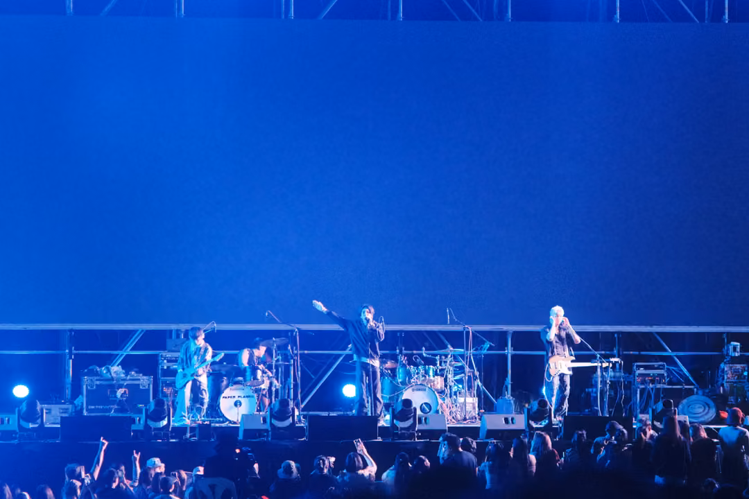 Band on a blue-lit stage, the singer raising one arm, crowd with raised hands in the foreground.