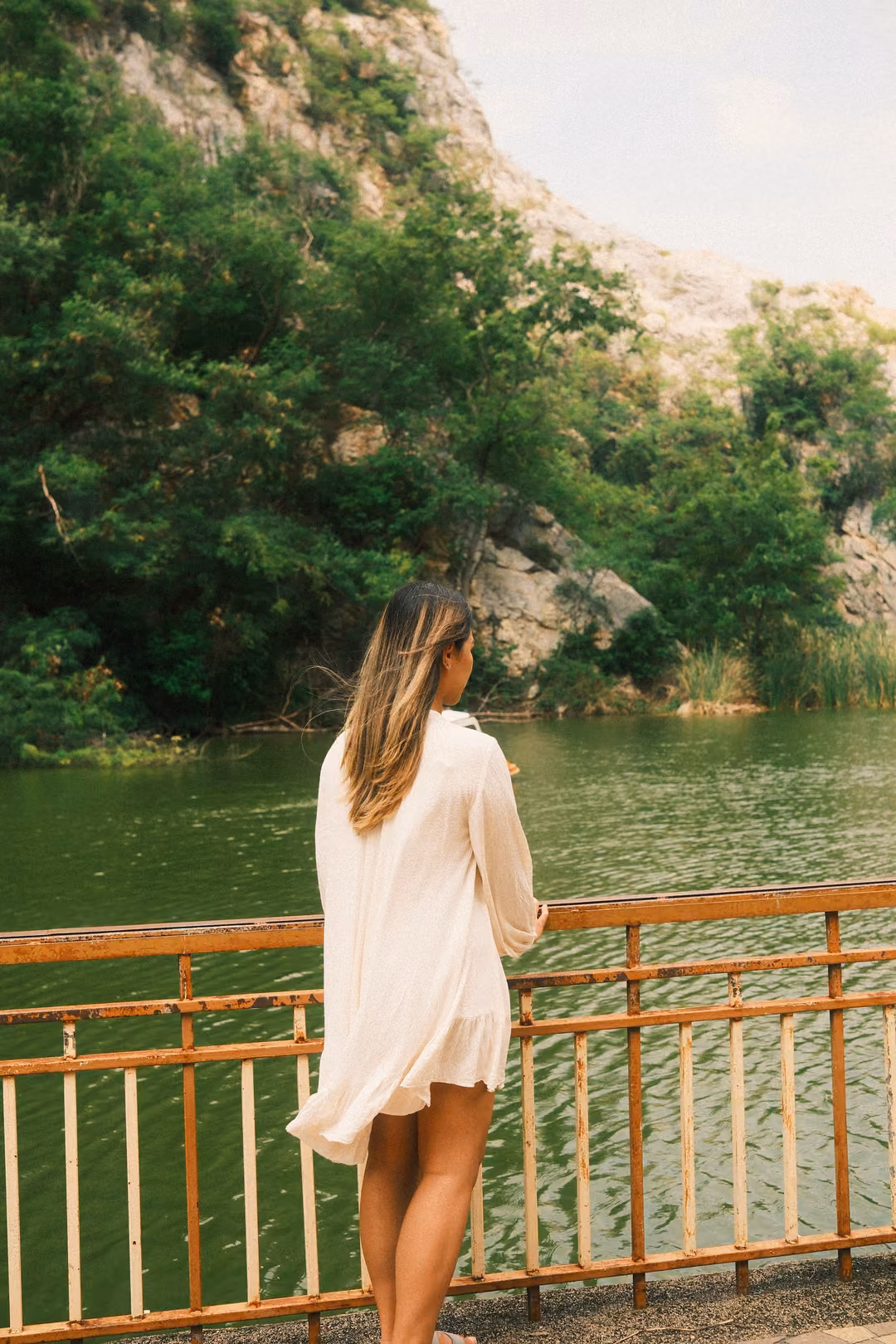 Woman in a white dress seen from behind, standing at a railing looking out over a green lake toward cliffs