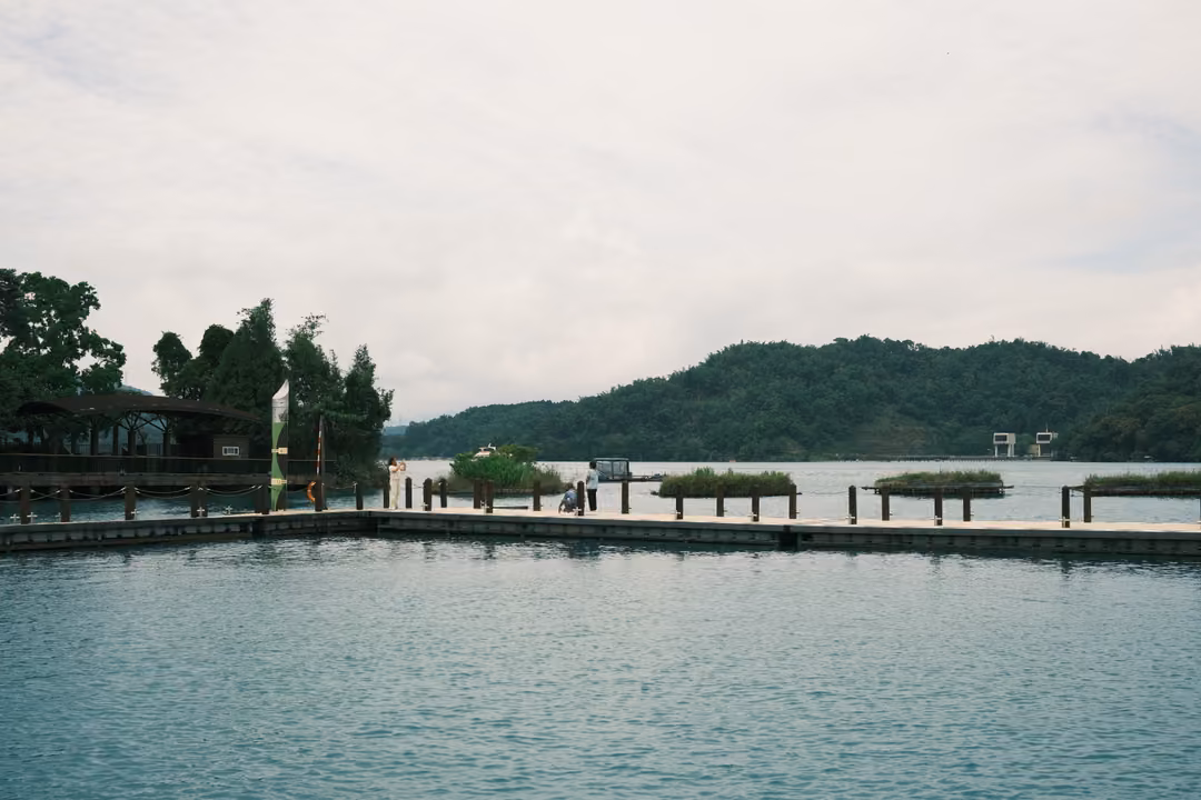 Floating wooden walkways and small planted rafts on a lake, a pavilion among trees and hills beyond