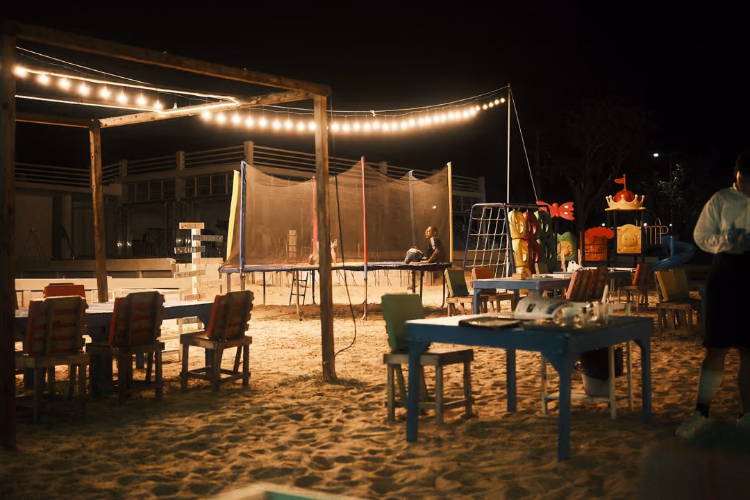 Beachside dining area at night under string lights, wooden chairs and blue tables on sand, a trampoline behind