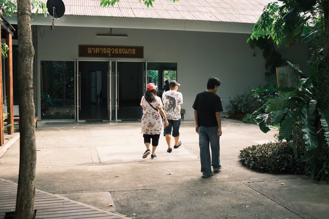 Three visitors walking toward a pale building with glass doors and a Thai sign, trees around