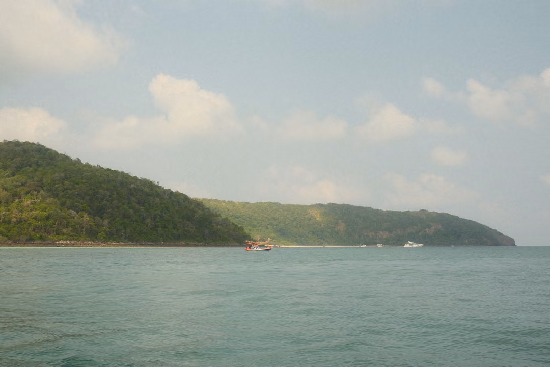 Two green headlands across a turquoise sea, a small red boat between them under a blue sky.