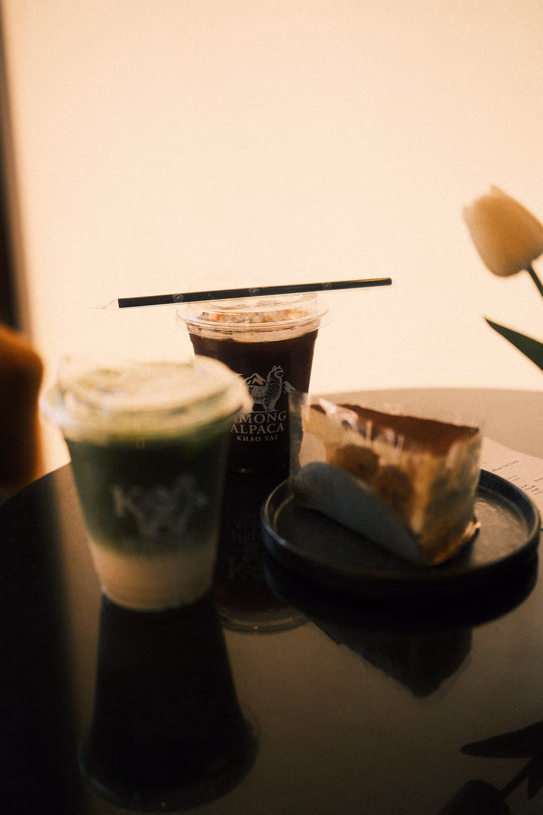 On a dark table, a green layered iced drink, an iced coffee with a straw, a cake slice and a white tulip.
