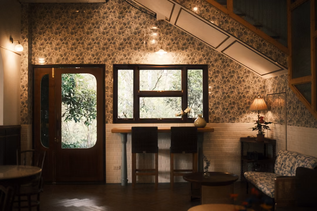 Floral wallpapered wall beneath a staircase with an arched glass door, window, bar stools, lamp and sofa