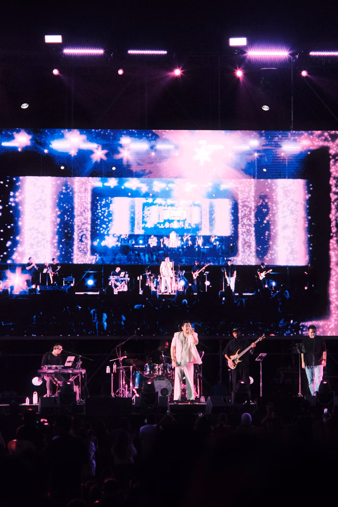 Stage beneath a screen of blue starbursts and pink sparkles framing a smaller image of the band
