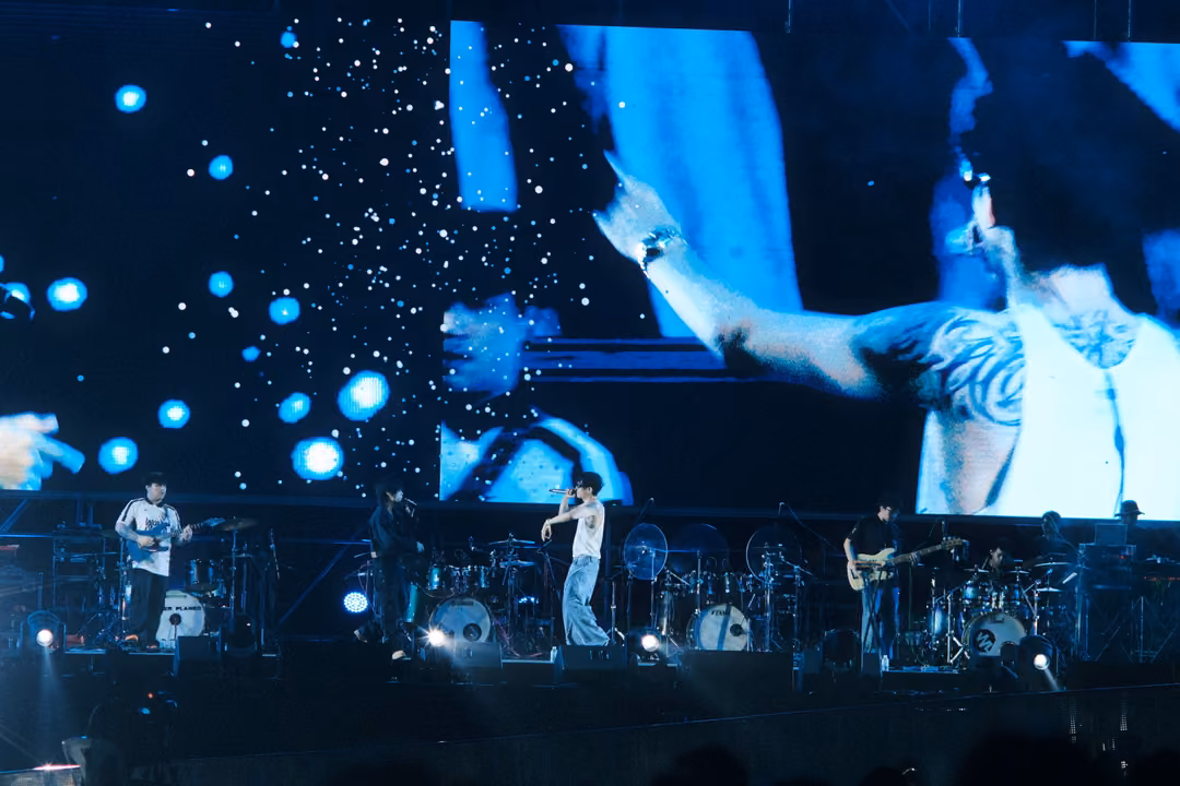 Screen showing the singer's tattooed arm reaching out, a watch on the wrist, band in blue light on the stage below.