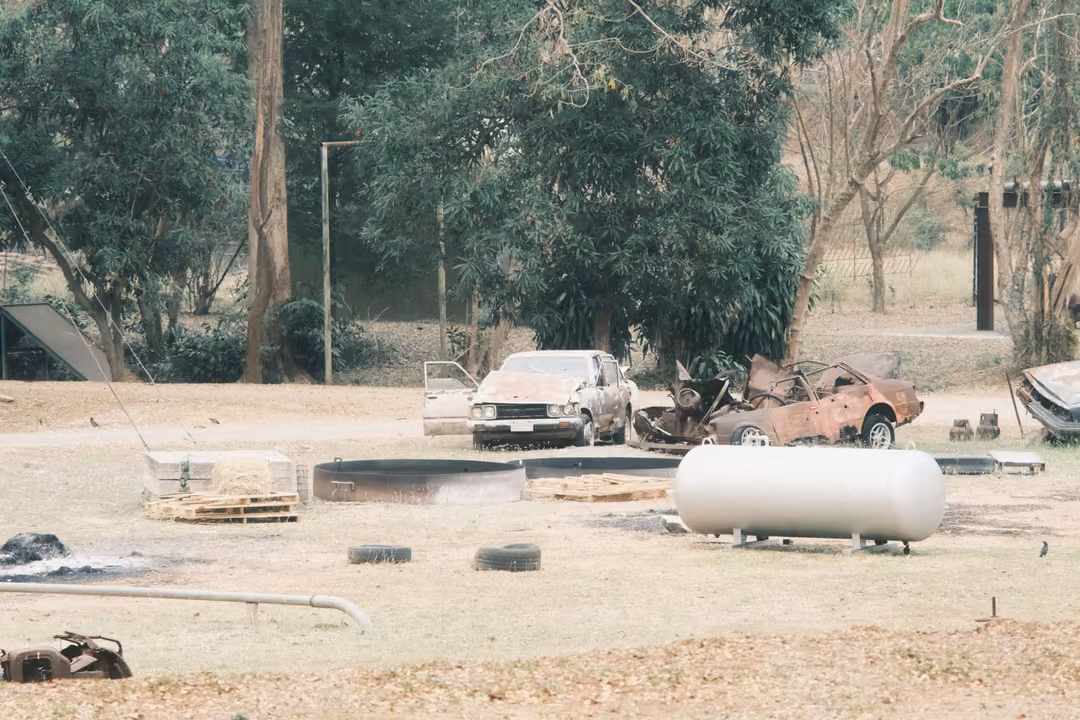 Dusty yard with wrecked cars, a white cylindrical tank, tyres and pallets under tall trees