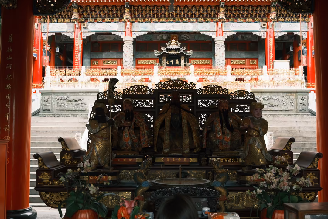 Temple hall altar with seated figures in front of a carved screen, a bright courtyard and pavilion beyond