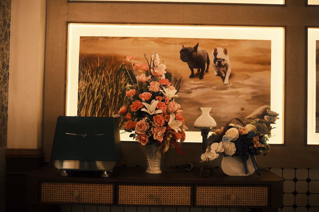 Lit painting of two small dogs on sand above a cabinet with flowers, a lamp and a green radio in dim light