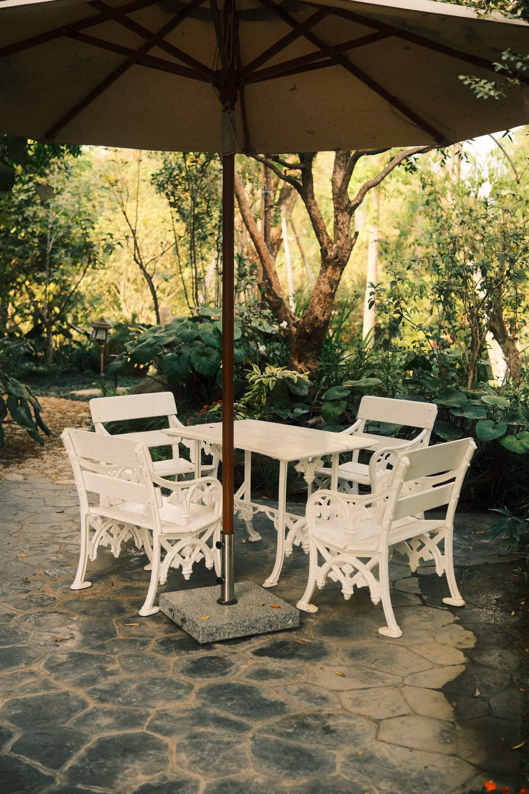 White ornate garden table and four chairs under a canvas umbrella on a stone patio among trees