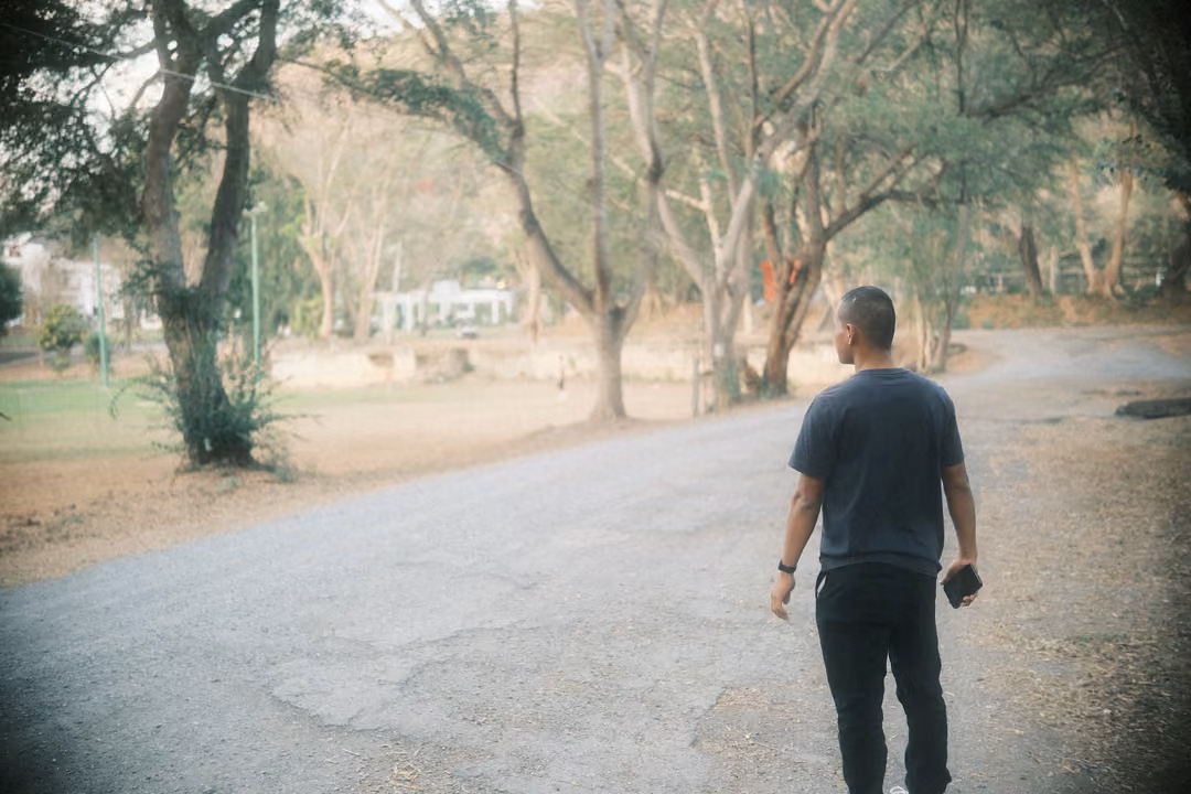 Man walking away down a cracked road toward dry trees and a field, holding a phone at his side