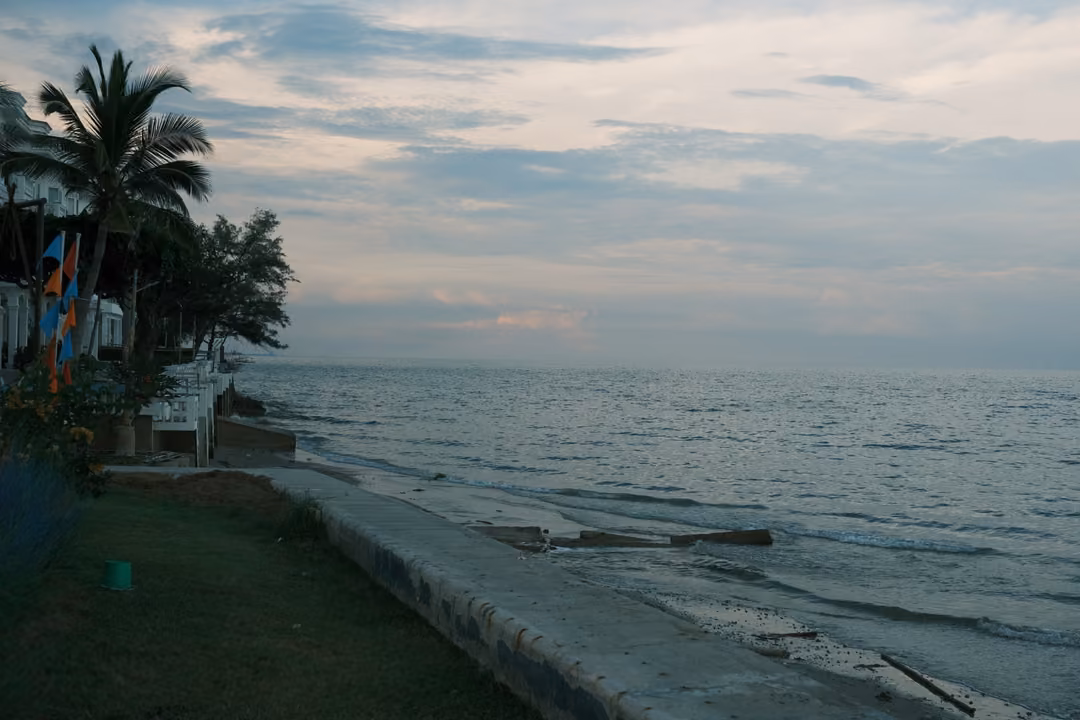 Seawall and grassy promenade along the shore, palms and colourful flags at the left, broken concrete in the water