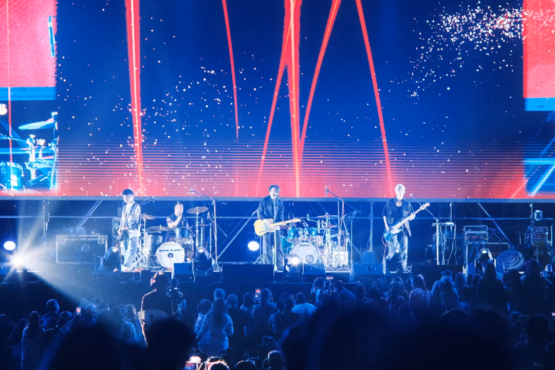 Band on stage beneath a large screen of red light beams and white sparkles on deep blue, crowd lit from the side