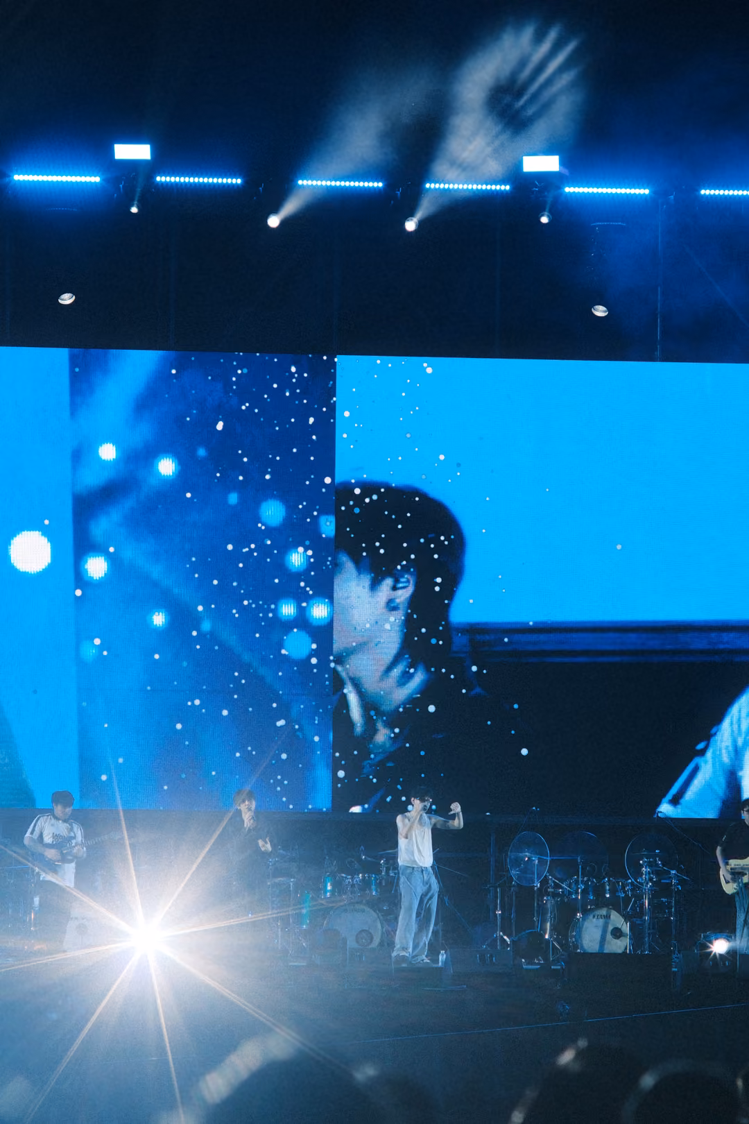 Vertical view of a blue stage with a bright starburst light at lower left and a screen close-up of a singer above.