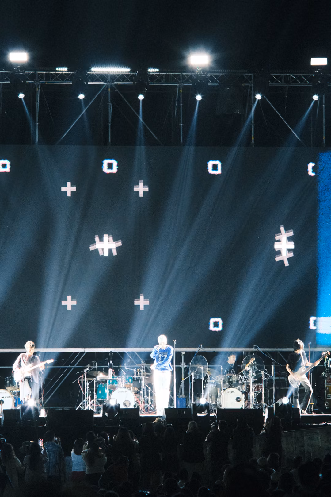 Vertical view of a screen of plus signs and hash shapes, white spotlights blazing, singer and guitarists on stage below.
