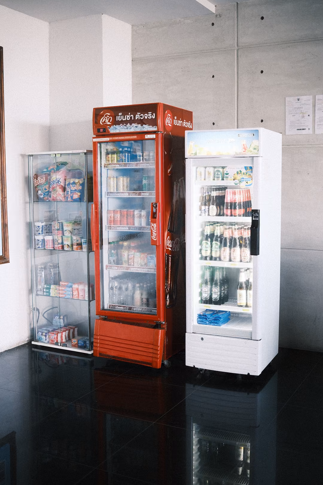 Three drink fridges, one red, one white and one glass display, against a grey concrete wall on a dark glossy floor