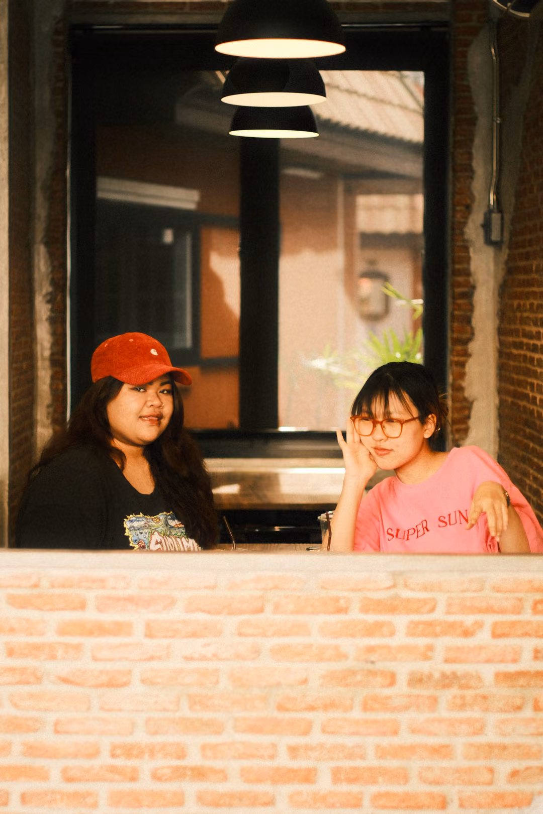 Two women posed behind a brick wall at a window, the one in pink pressing a hand to her cheek