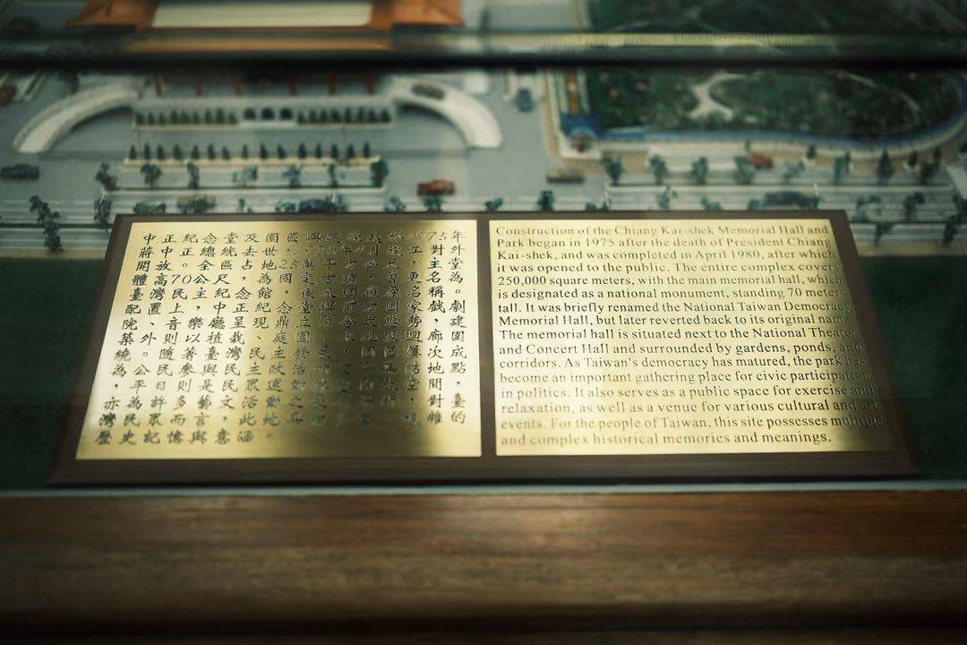 Brass plaque with Chinese and English text set before a scale model of a large memorial complex under glass