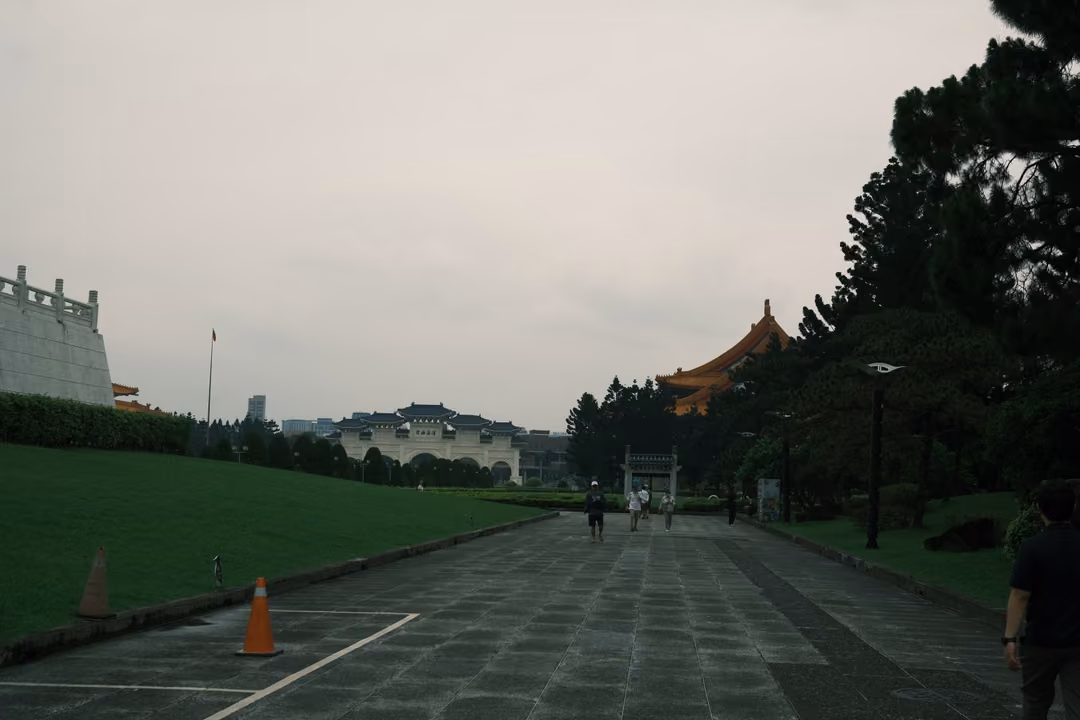 Broad paved avenue between lawns and pine trees leading toward a distant gate and orange-roofed building