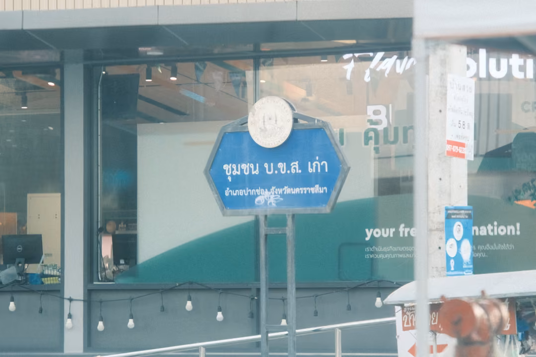 A blue hexagonal Thai street sign on posts in front of a glass shop window with string lights and green signage.