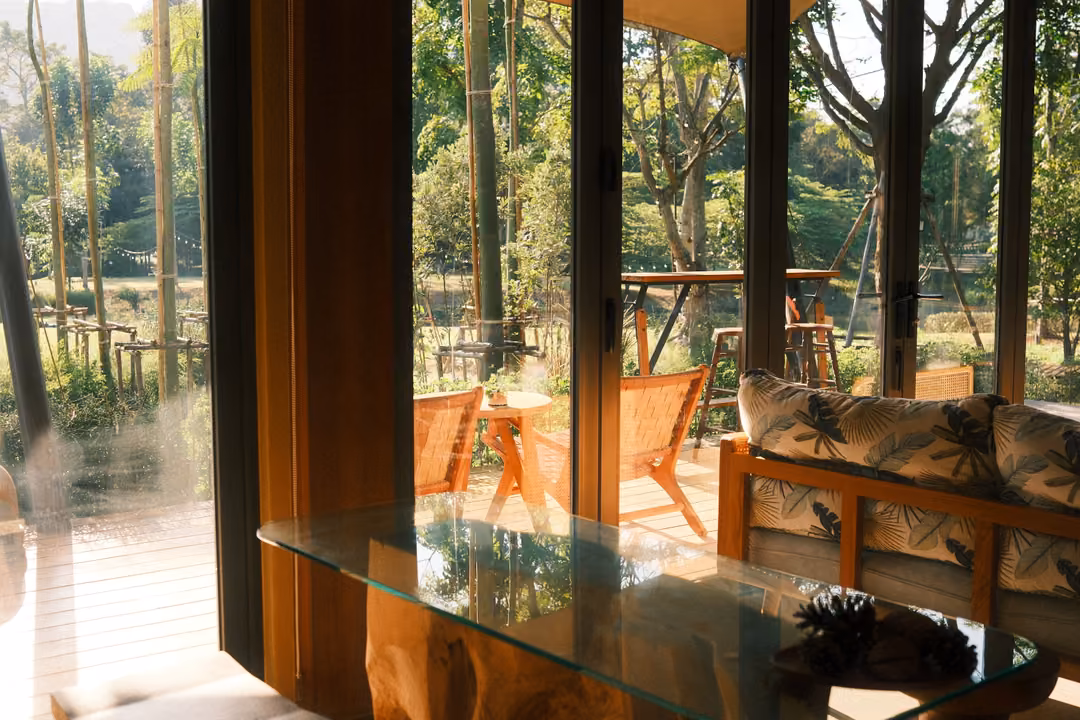 Glass-topped table and cushioned sofa beside tall windows, wooden chairs on a sunlit terrace outside