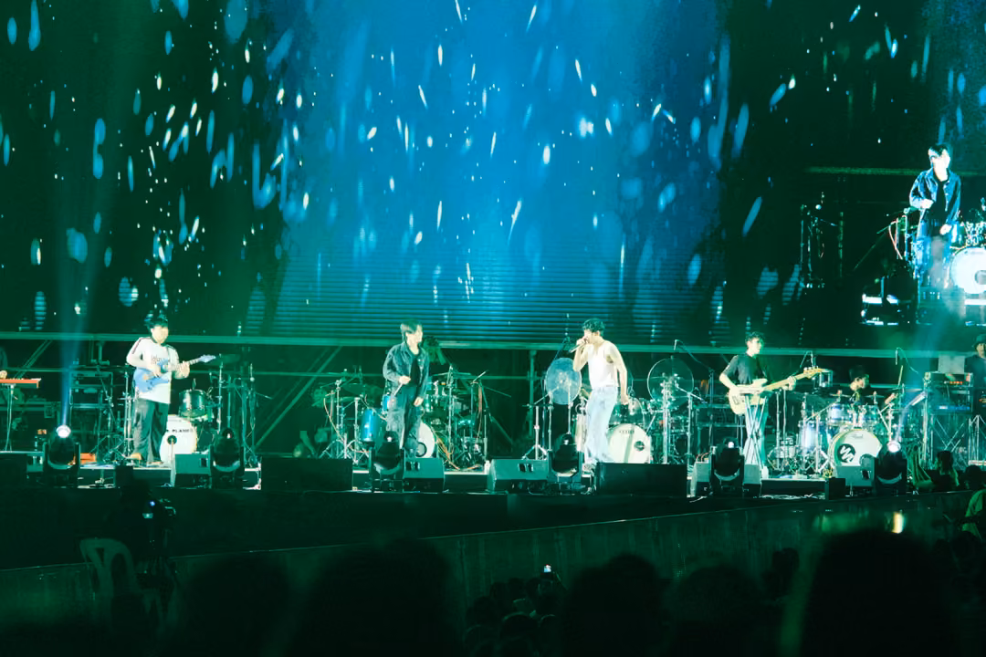 Band performing beneath a blue-green screen of falling white flakes, a drummer visible high at right.
