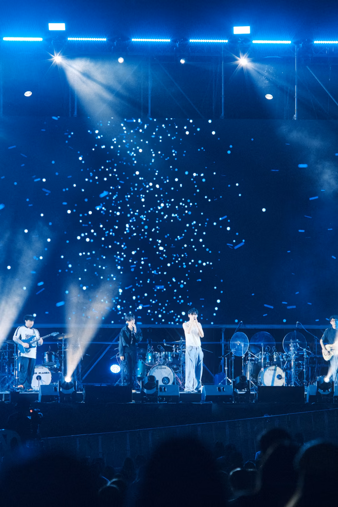 Stage at night with spotlights blazing from the truss into a screen of white flecks, band in a line below.