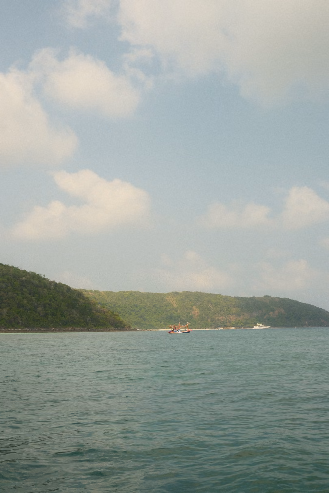 Green hills over a blue-green sea, small boats moored along the shore under a blue sky with clouds.