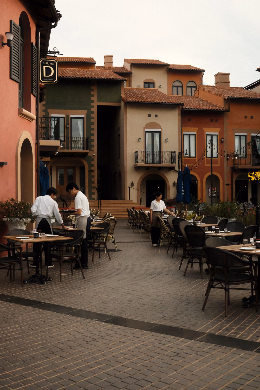 Waiters setting tables at an outdoor café in a paved square framed by pastel Italian-style buildings