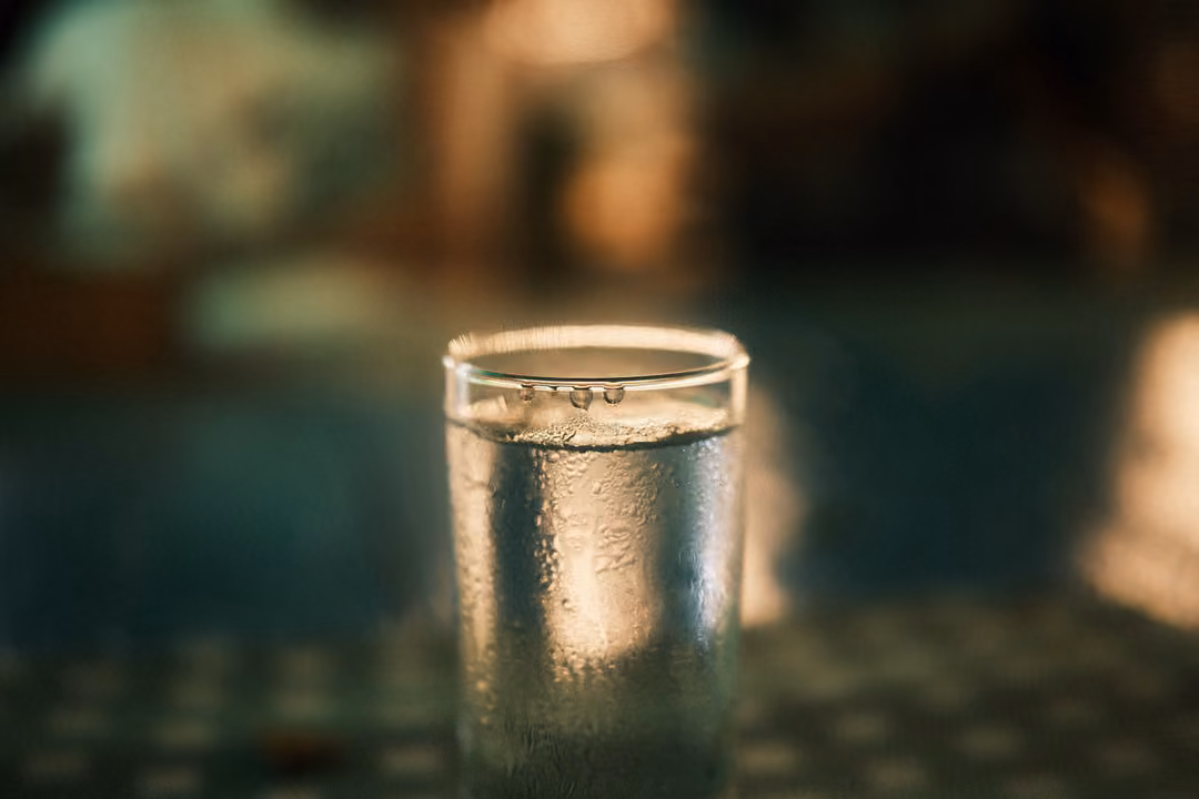 A cold glass of water with condensation on a table at night, blurred lights and a pool behind