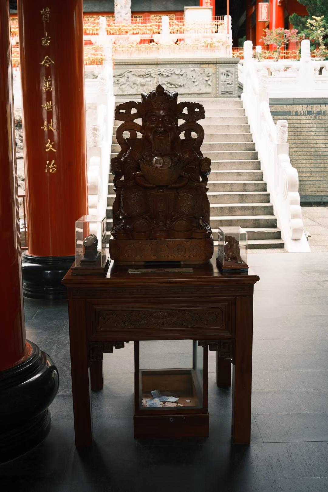 Carved wooden throne-like chair on a table in a dim temple hall, red pillars and white stairs behind
