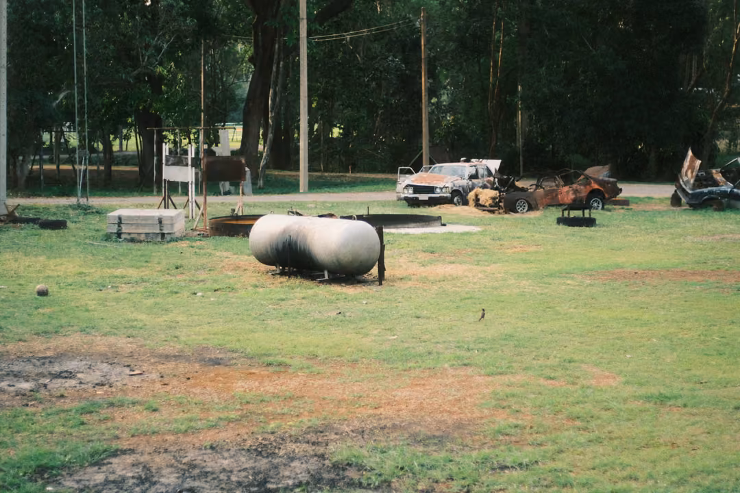 A grassy field with a white gas tank, burnt-out cars, poles and ropes, trees in the background.