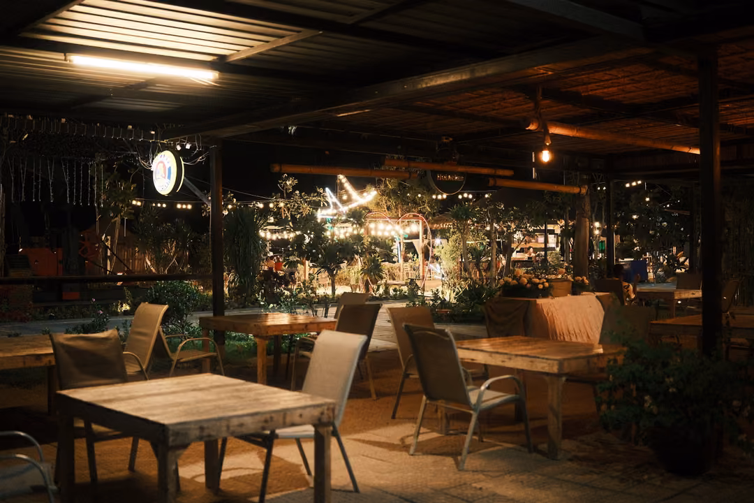 Open-sided restaurant at night with wooden tables and chairs under a corrugated roof, plants and fairy lights beyond