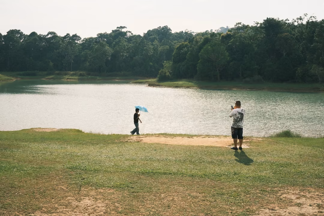 One person holds a pale blue umbrella by a lake while another in a white T-shirt takes a phone photo.