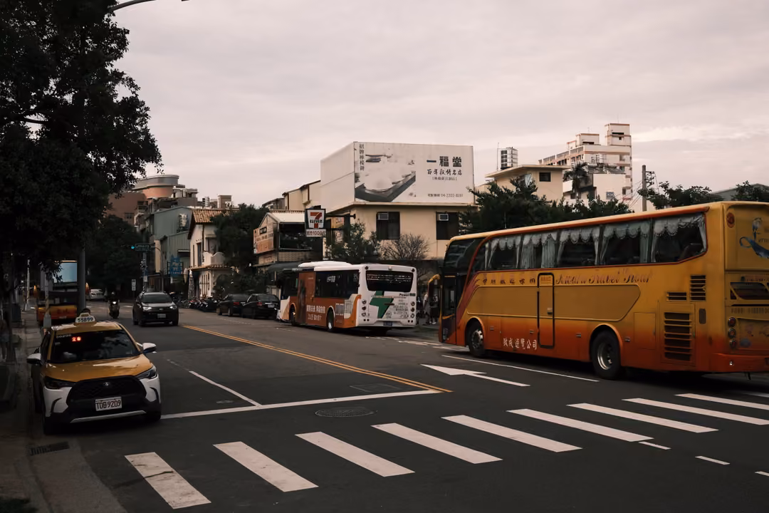 Yellow coach bus and a taxi on a city street beneath an overcast sky, crosswalk stripes in front