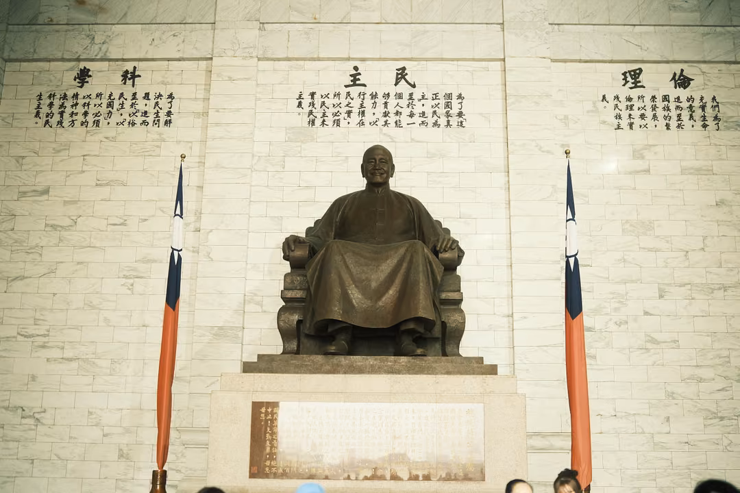 Seated bronze statue on a stone plinth against pale marble wall with carved characters and two flags