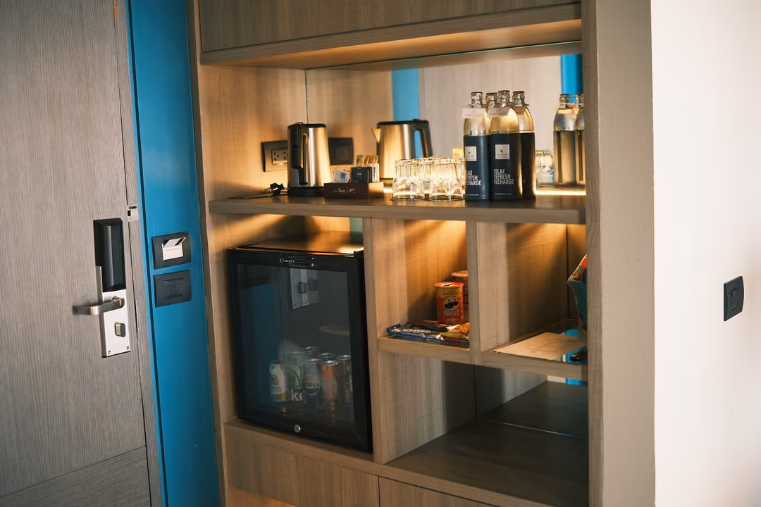 A hotel room shelf with a kettle, bottled water, glasses and a mini fridge beside the door lock
